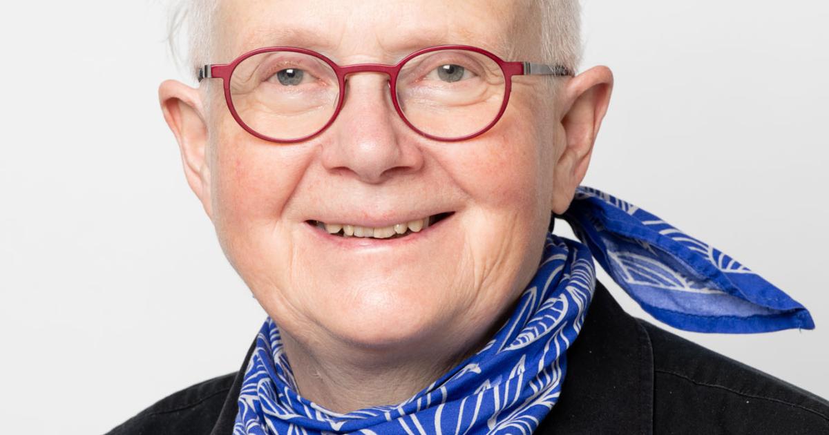 Portrait of artist Rita McKeough. She has short white hair, glasses, and is wearing a blue scarf and dark jean jacket.