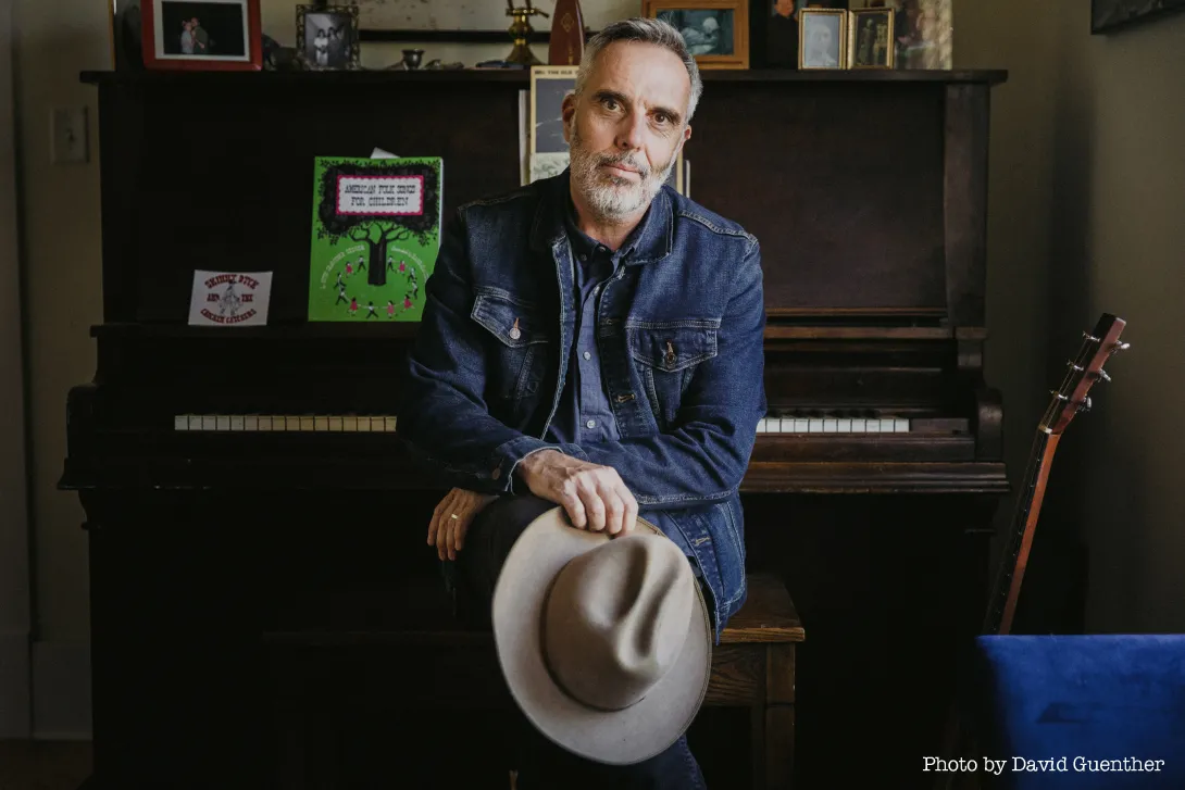 Man with short white beard in jean jacket an holding white cowboy hat leans against an upright piano